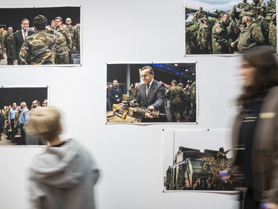 Fünf Fotos vom Verteidigungsminister hängen an einer weißen Wand, zwei Personen laufen an den Bildern vorbei