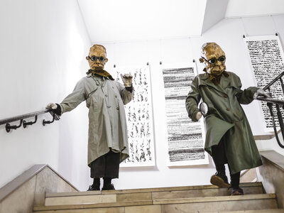 Zwei Personen in Trenchcoats und Masken wandern durch das Treppenhaus